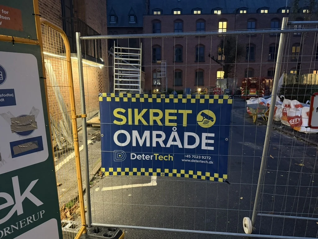 Lille blåt og gult DeterTech-banner med SIKRET OMRÅDE og kameraikon på byggepladshegn