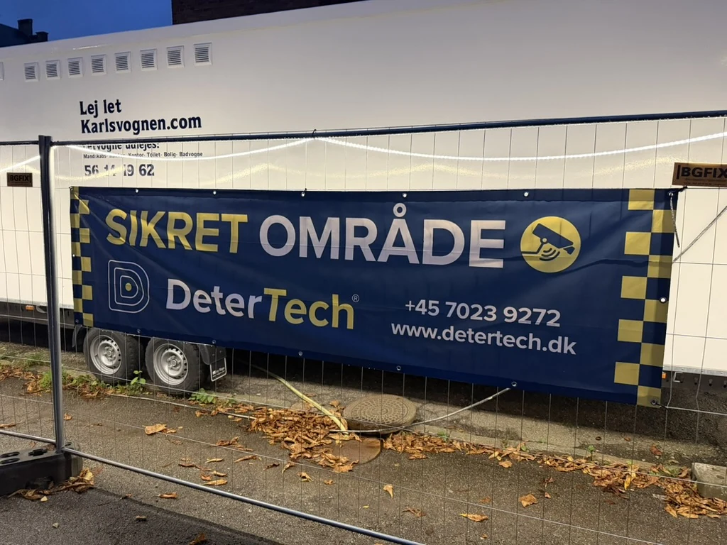 Nærbillede af bredt DeterTech-banner med kantforstærkning og øjer på hegn foran skurvogn